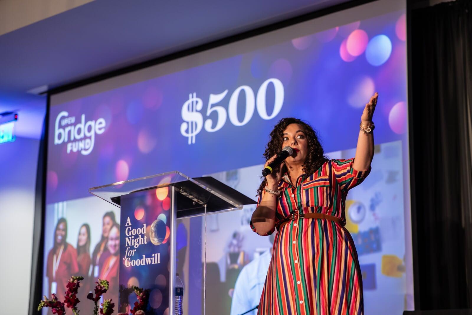 Micki Pickering in a multicolored dress, hosting as event emcee at a podium with a bid screen behind her, indicating a $500 paddle raise level